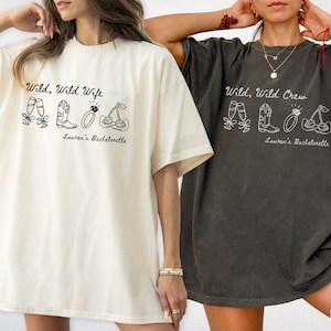 Puede incluir: Dos camisetas holgadas, una marfil y otra color pimienta, presentan los textos "Wild, Wild Wife" y "Wild, Wild Crew" con ilustraciones de copas de champ&aacute;n, botas de vaquero, un anillo y cerezas. Tambi&eacute;n se incluye el texto "Lauren's Bachelorette".