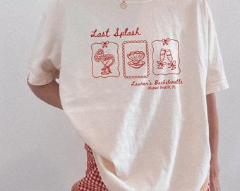 May include: Ivory-colored t-shirt with red graphic text and illustrations. The text reads "Last Splash" above three framed images: a mermaid cocktail, a seashell, and champagne flutes. Below, it says "Lauren's Bachelorette Miami Beach, FL."