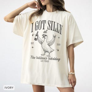 Op de afbeelding: Ivoor kleurig t-shirt met een afbeelding van een eend met een zonnebril en een fles. De tekst luidt "I GOT SILLY at The Wilson's Wedding LAS VEGAS" met het jaar 2026 en "EST." op het shirt.