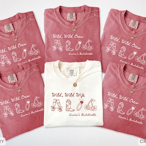 Puede incluir: Una colecci&oacute;n de camisetas carmes&iacute; y marfil con las frases "Wild, Wild Crew" y "Wild, Wild Wife". Cada camiseta presenta un collar y un gr&aacute;fico de copas de champ&aacute;n, botas, un anillo y cerezas. Son camisetas para una despedida de soltera.