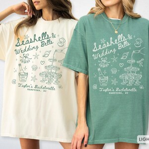 K&ouml;nnte beinhalten: Zwei T-Shirts, eines in Elfenbein und eines in Hellgr&uuml;n, mit dem Text "Seashells & Wedding Bells" und "Taylor's Bachelorette, Hamptons, NY". Das Design umfasst Sektgl&auml;ser, Muscheln und eine Sektflasche.