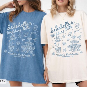 K&ouml;nnte beinhalten: Zwei T-Shirts, eines in Jeansblau und eines in Elfenbein, mit einem grafischen Design. Das Design enth&auml;lt den Text "Seashells & Wedding Bells" und "Taylor's Bachelorette, Hamptons, NY" sowie Muscheln, Sektgl&auml;ser und eine Flasche.