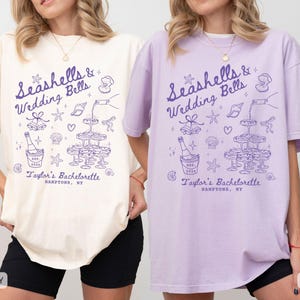K&ouml;nnte beinhalten: Elfenbein- und orchideenfarbene T-Shirts mit einem lila Grafikdesign. Das Design enth&auml;lt den Text "Seashells & Wedding Bells" und "Taylor's Bachelorette, Hamptons, NY" sowie Illustrationen von Sektgl&auml;sern, Muscheln und einer Sektflasche.