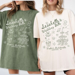 K&ouml;nnte beinhalten: Zwei &uuml;bergro&szlig;e T-Shirts, eines in Moosgr&uuml;n und eines in Elfenbein, mit einer wei&szlig;en Grafik. Die Grafik zeigt den Text "Seashells & Wedding Bells" mit Illustrationen von Champagner, Muscheln und Sektgl&auml;sern. Der Text "Taylor's Bachelorette, Hamptons, NY" ist ebenfalls enthalten.