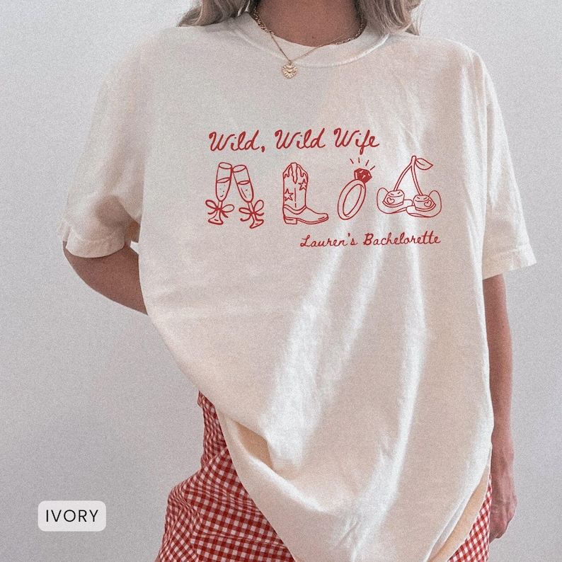 Puede incluir: Camiseta color marfil con la frase "Wild, Wild Wife" en rojo, junto con ilustraciones de copas de champ&aacute;n, una bota de vaquero, un anillo y cerezas. El texto "Lauren's Bachelorette" tambi&eacute;n est&aacute; impreso en la camiseta.