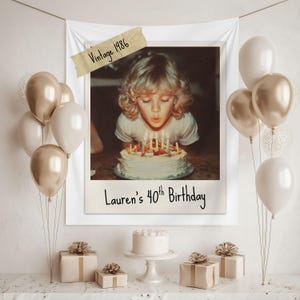 Op de afbeelding: Een verjaardagsscène in vintage stijl. Een ingelijste foto toont een persoon die kaarsen uitblaast op een taart. De foto is gelabeld met "Vintage 1986" en "Lauren's 40th Birthday". Gouden en witte ballonnen, cadeaus en een kleine taart maken de feestelijke decoratie compleet.