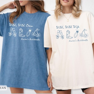 Puede incluir: Dos camisetas holgadas, una azul vaquero y otra marfil, con los textos "Wild, Wild Crew" y "Wild, Wild Wife". Cada camiseta presenta un gr&aacute;fico blanco de copas de champ&aacute;n, una bota de vaquero, un anillo y cerezas. "Lauren's Bachelorette" est&aacute; impreso debajo.