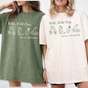 Puede incluir: Dos camisetas holgadas, una verde musgo y otra crema, con los textos "Wild, Wild Crew" y "Wild, Wild Wife" en letra cursiva. Cada camiseta presenta dibujos de zapatillas de ballet, botas de vaquero, un anillo y cerezas. "Lauren's Bachelorette" est&aacute; impreso debajo.