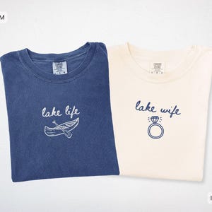 May include: Two t-shirts, one denim blue and one ivory. The denim shirt has "lake life" with a canoe graphic. The ivory shirt says "lake wife" with a diamond ring graphic. Both shirts have a crew neck.