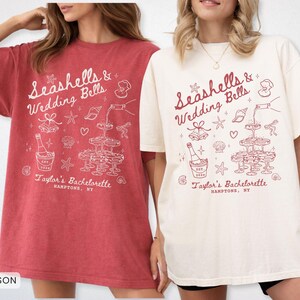 K&ouml;nnte beinhalten: Zwei &uuml;bergro&szlig;e T-Shirts, eines in Karmesinrot und eines in Elfenbein, mit einem grafischen Design. Das Design enth&auml;lt den Text "Seashells & Wedding Bells" und "Taylor's Bachelorette, Hamptons, NY" mit Illustrationen.