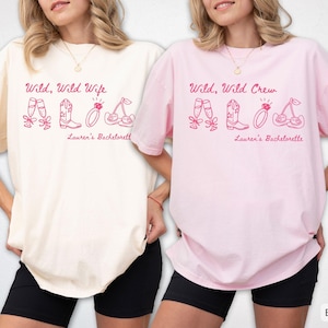 Puede incluir: Dos camisetas, una marfil y otra rosa, con el texto "Wild, Wild Wife" y "Wild, Wild Crew" en escritura rosa. Cada camiseta tiene ilustraciones a juego de copas de champ&aacute;n, botas, un anillo y cerezas. El texto "Lauren's Bachelorette" est&aacute; debajo.