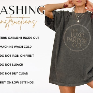 K&ouml;nnte beinhalten: Dunkelgraues T-Shirt mit rundem Logo "The Luxe Party Co." und Waschanleitung. Die Anweisungen umfassen das Wenden des Kleidungsst&uuml;cks, Maschinenw&auml;sche kalt und Trocknen bei niedriger Temperatur.
