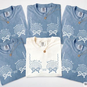 Puede incluir: Varios camisetas azul vaquero y marfil con un diseño de concha y el texto "Taylor's Back, Hamptons, NY 2026". Cada camiseta tiene un collar dorado con un colgante redondo. Las camisetas están dispuestas sobre un fondo blanco.