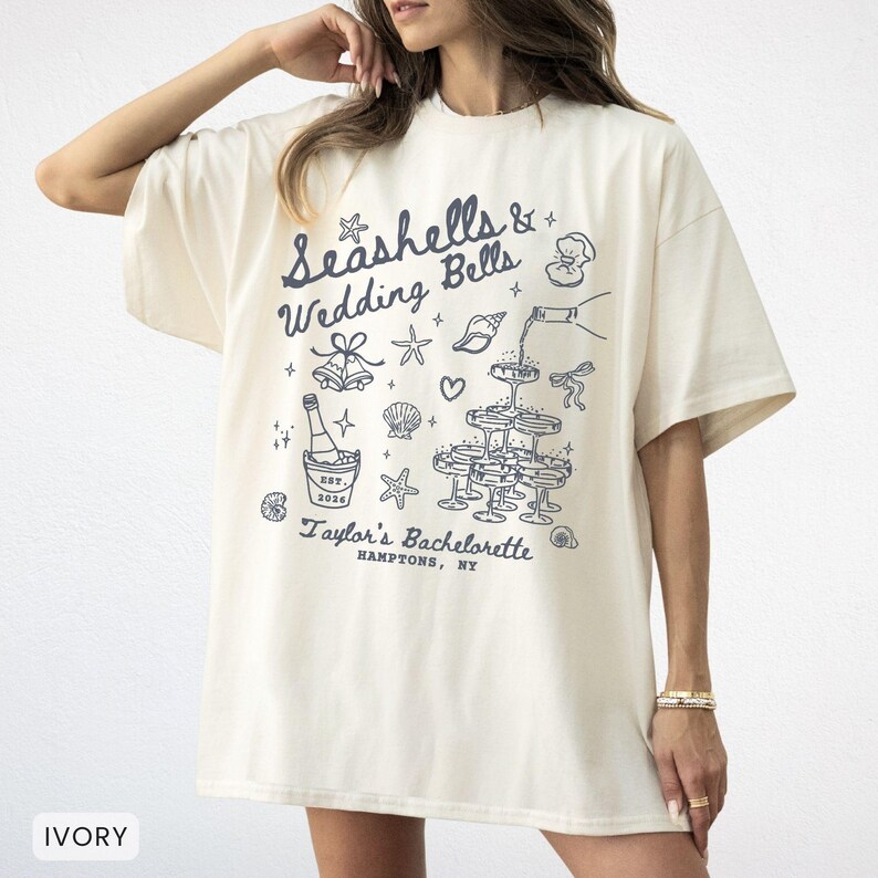 K&ouml;nnte beinhalten: Elfenbeinfarbenes T-Shirt mit einem marineblauen Aufdruck, der "Seashells & Wedding Bells" und "Taylor's Bachelorette, Hamptons, NY" zeigt. Das Design umfasst Illustrationen von Muscheln, Champagner und Sektgl&auml;sern.