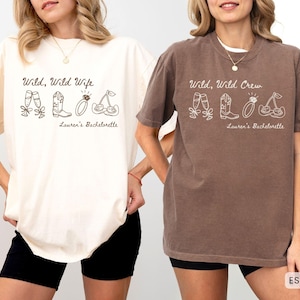 Puede incluir: Dos camisetas, una marfil y otra espresso, con el texto "Wild, Wild Wife" y "Wild, Wild Crew" en letra cursiva. Cada camiseta tiene un dibujo lineal de zapatillas de ballet, botas de vaquero, un anillo y cerezas. "Lauren's Bachelorette" est&aacute; debajo.