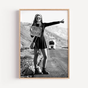 Könnte beinhalten: Ein Schwarz-Weiß-Foto einer Person in Nonnentracht, Sonnenbrille und kurzem Kleid, die am Straßenrand steht und per Anhalter fährt. Die Person hält ein Schild mit der Aufschrift TO HEAVEN und trägt eine Umhängetasche sowie eine große Reisetasche.