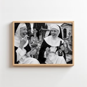 May include: A black and white framed photograph depicting two individuals dressed in traditional nun habits, laughing in a cemetery. One holds a lit cigarette, while the other holds a dark glass bottle with a label reading Holy Grail.