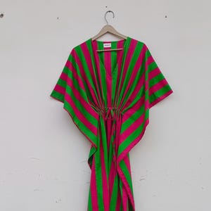 May include: A vibrant, striped kaftan in shades of hot pink and green hangs from a wooden hanger. The lightweight fabric features bold vertical stripes and a drawstring waist. This flowing garment is perfect for beachwear or casual wear.