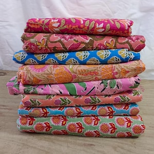 Op de afbeelding: Een stapel gevouwen stoffen met bloemmotieven in verschillende kleuren, waaronder roze, blauw, oranje en groen. De stoffen zijn gestapeld op een houten oppervlak, met een witte achtergrond.