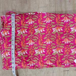 May include: A folded piece of pink fabric with a repeating floral pattern in white and orange. A measuring tape is placed along the edge. The fabric has a tag that reads "TEXTILECITY store".