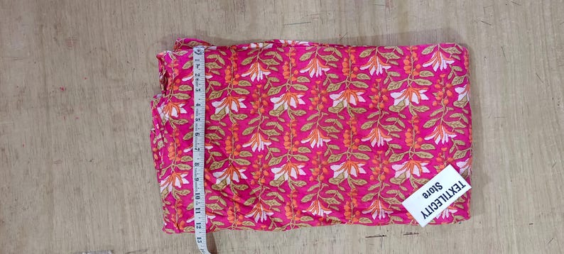 May include: A folded piece of pink fabric with a repeating floral pattern in white and orange. A measuring tape is placed along the edge. The fabric has a tag that reads "TEXTILECITY store".