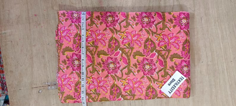 May include: Pink fabric with a floral pattern in shades of pink and brown. A measuring tape shows the fabric is approximately 14 inches long. The tag reads TEXTILECITY Store.