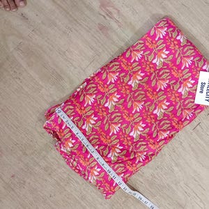 May include: A folded piece of pink fabric with a repeating floral pattern in white and orange. A measuring tape is laid across the fabric. The tag reads TEXTILECITY Store.