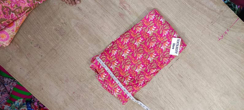May include: A folded piece of pink fabric with a repeating floral pattern in white and orange. A measuring tape is laid across the fabric. The tag reads TEXTILECITY Store.