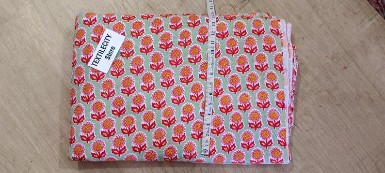 May include: A folded fabric with a repeating floral pattern in red, orange, and white on a light green background. A measuring tape is laid across the fabric. The tag says TEXTILECITY Store.