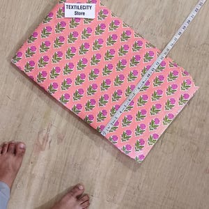 May include: A folded fabric with a repeating floral pattern in pink and green. A measuring tape is laid across the fabric. The text "TEXTILECITY Store" is on a label on the fabric.