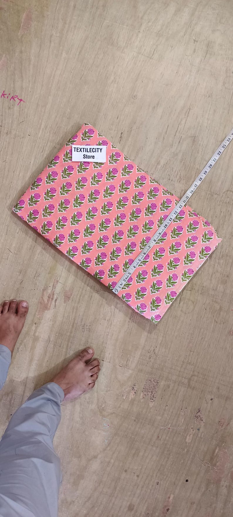May include: A folded fabric with a repeating floral pattern in pink and green. A measuring tape is laid across the fabric. The text "TEXTILECITY Store" is on a label on the fabric.