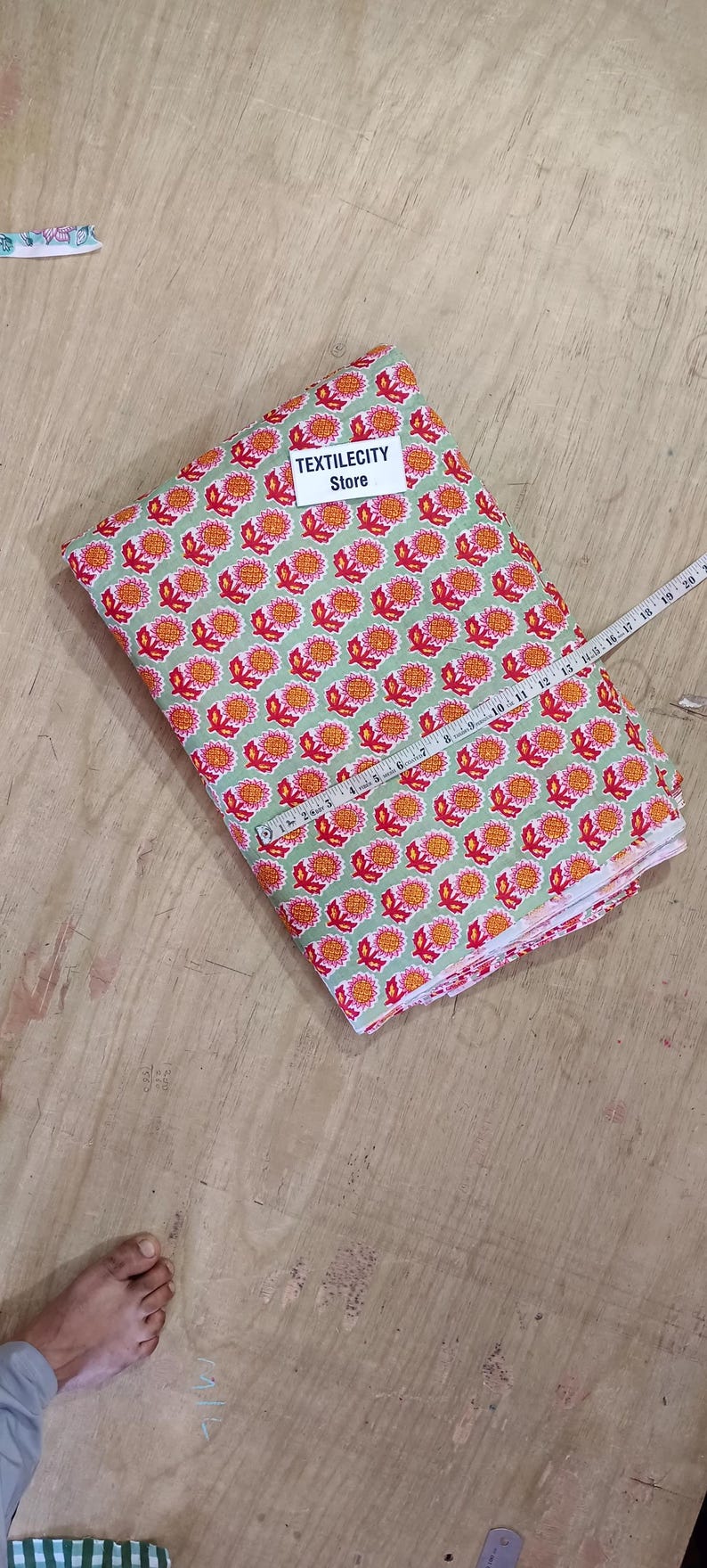 May include: A folded fabric with a repeating floral pattern in red and orange on a light green background. A measuring tape is laid across the fabric. The label "TEXTILECITY Store" is visible.