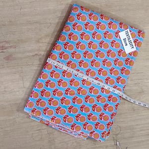 May include: A folded fabric with a repeating orange fruit pattern on a light blue background. A white measuring tape is laid across the fabric. A tag that says "TEXTILECITY Store" is attached to the fabric.