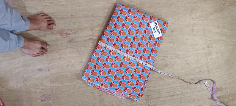 May include: A folded fabric with a repeating orange fruit pattern on a light blue background. A white measuring tape is laid across the fabric. A tag that says "TEXTILECITY Store" is attached to the fabric.