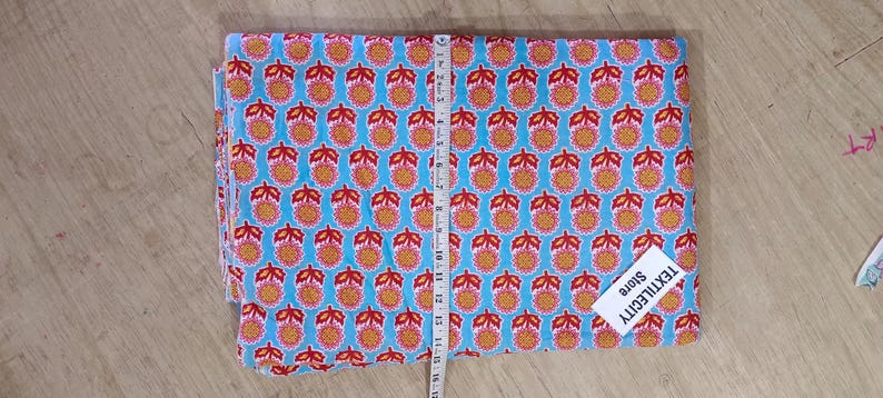 May include: Folded fabric with a repeating pattern of orange and red floral designs on a light blue background. A measuring tape is placed on top of the fabric. The tag says TEXTILECITY Store.