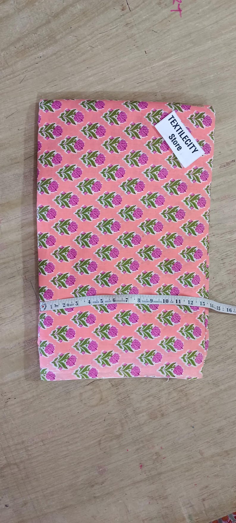 May include: Pink fabric with a repeating floral pattern in pink and green. A measuring tape is laid across the fabric. The tag reads TEXTILECITY Store.