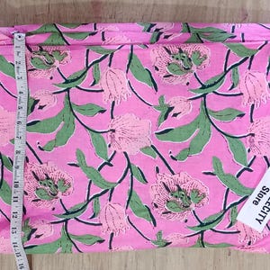 May include: Pink floral fabric with a green and pink flower and leaf pattern. A measuring tape is placed along the edge of the fabric. The fabric has a tag that says TEXTILECITY Store.