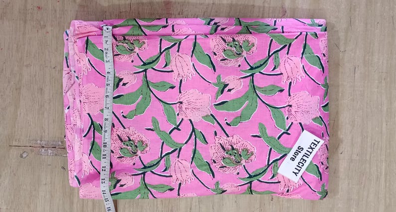 May include: Pink floral fabric with a green and pink flower and leaf pattern. A measuring tape is placed along the edge of the fabric. The fabric has a tag that says TEXTILECITY Store.