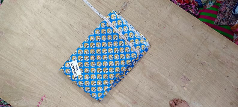 May include: A folded blue fabric with a repeating yellow floral pattern, measured with a white measuring tape. The fabric has a tag that reads "TEXTILECITY STORE". The fabric is on a light brown wooden surface.