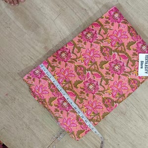 May include: Pink floral fabric with a tape measure laid across it. The fabric has a repeating pattern of pink flowers and green leaves on a peach background. The fabric is labeled "TEXTILECITY Store".