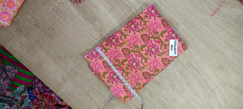May include: Pink floral fabric with a tape measure laid across it. The fabric has a repeating pattern of pink flowers and green leaves on a peach background. The fabric is labeled "TEXTILECITY Store".