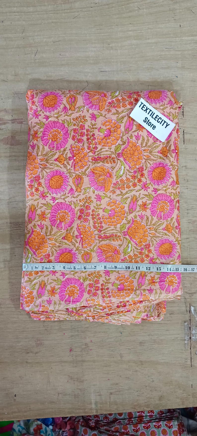 May include: A folded fabric with a floral pattern in shades of pink, orange, and green. The fabric is measured with a tape measure. The tag says TEXTILECITY Store.