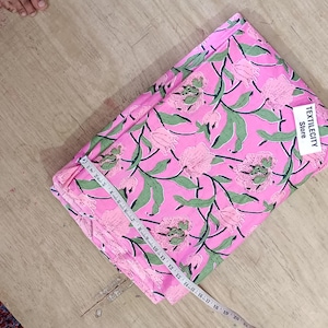 May include: Folded pink fabric with a floral pattern of pink flowers and green leaves. A white tag with the text "TEXTILEGITY Store" is attached. A measuring tape is placed across the fabric. The fabric is on a wooden surface.