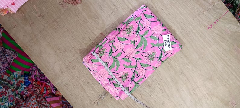 May include: Folded pink fabric with a floral pattern of pink flowers and green leaves. A white tag with the text "TEXTILEGITY Store" is attached. A measuring tape is placed across the fabric. The fabric is on a wooden surface.