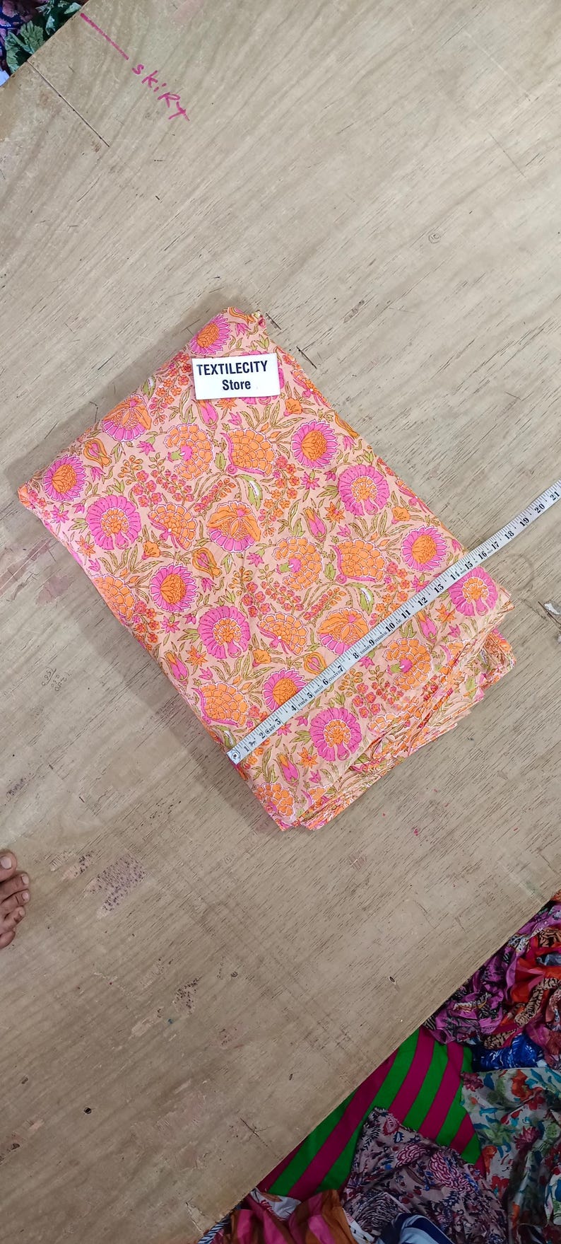 May include: A folded fabric with a floral pattern in shades of pink and orange. The fabric is being measured with a ruler. The text "TEXTILECITY Store" is visible on a white label.