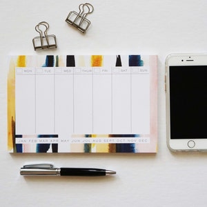 May include: A weekly planner with a colorful watercolor design, featuring sections for each day of the week and months of the year. Includes a smartphone, a pen, and two binder clips.