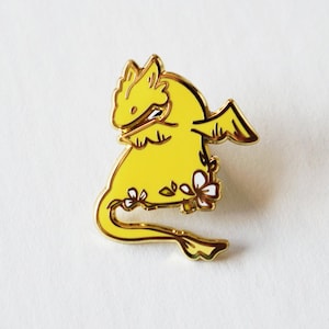May include: A yellow dragon enamel pin with gold trim. The dragon has small wings and is decorated with small white and pink flowers. The pin is set against a white background.