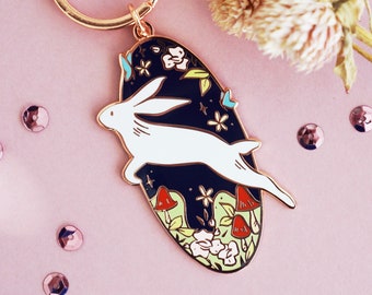 May include: A gold-colored keychain with a black and white enamel pendant featuring a white rabbit leaping across a dark blue background with flowers and mushrooms. The keychain is on a pink surface with sequins and dried flowers.