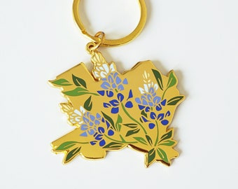 May include: Gold-colored keychain featuring a floral design of bluebonnets. The keychain has a gold ring and a charm shaped like a state outline. The bluebonnet flowers are detailed with blue, white, and green accents.