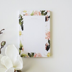 May include: A rectangular notepad with a floral design. The notepad has a white background with a border of pink roses, green leaves, and white flowers. The word "NOTES" is printed at the top.